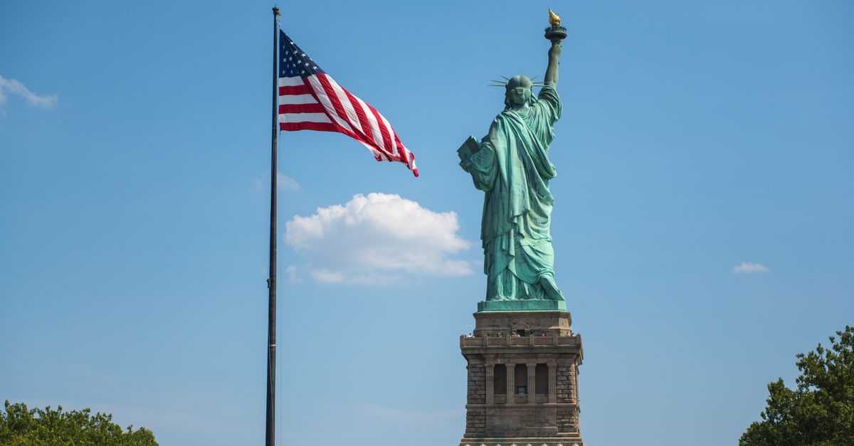 The Statue of Liberty alongside the American flag under a clear blue sky in New York City.